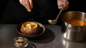 appetiser. magazine 6 Chef garnishing clam chowder served in a hollowed sourdough bread bowl, with a pot of chowder and a bowl of clams on a stainless steel counter.