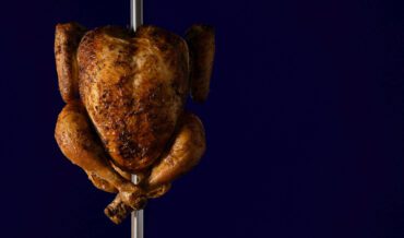 Whole rotisserie chicken cooked golden brown on a spit against a dark blue background.