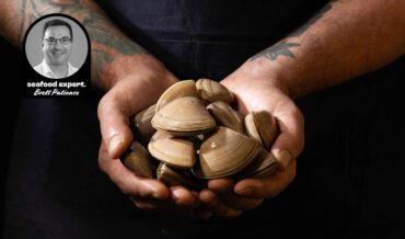 Tattooed hands holding a large handful of fresh Cloudy Bay clams, with a photo inset of seafood expert Brett Patience.