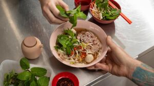 A bowl of bún bò Huế with round rice noodles, sliced beef, meatballs, bean sprouts, fresh mint and coriander, garnished with chilli and served with herbs on the side.
