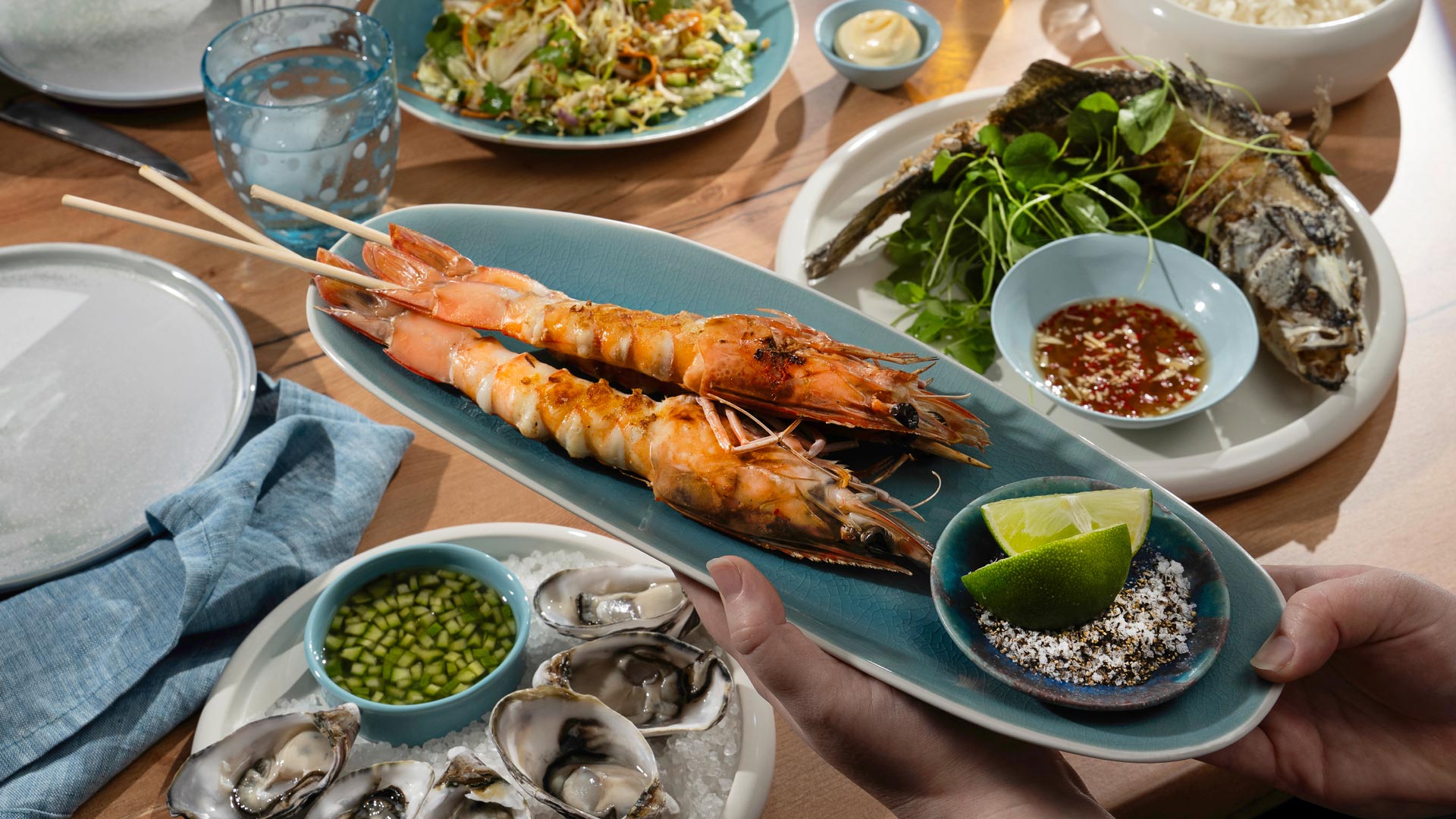 Grilled prawns on skewers served on a blue platter with lime wedges and a dish of salt and pepper for muối tiêu chanh. Surrounding dishes include oysters with cucumber dipping sauce, fried whole fish with herbs and nước chấm, and a fresh salad.
