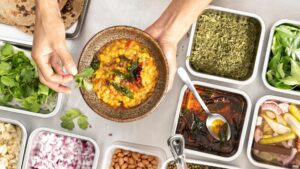 appetiser. magazine 8 A top-down view of a person holding a bowl of food, known as Indian Dal Tadka. The dish is garnished with fresh herbs and spices, surrounded by various ingredient containers on a rustic wooden surface.