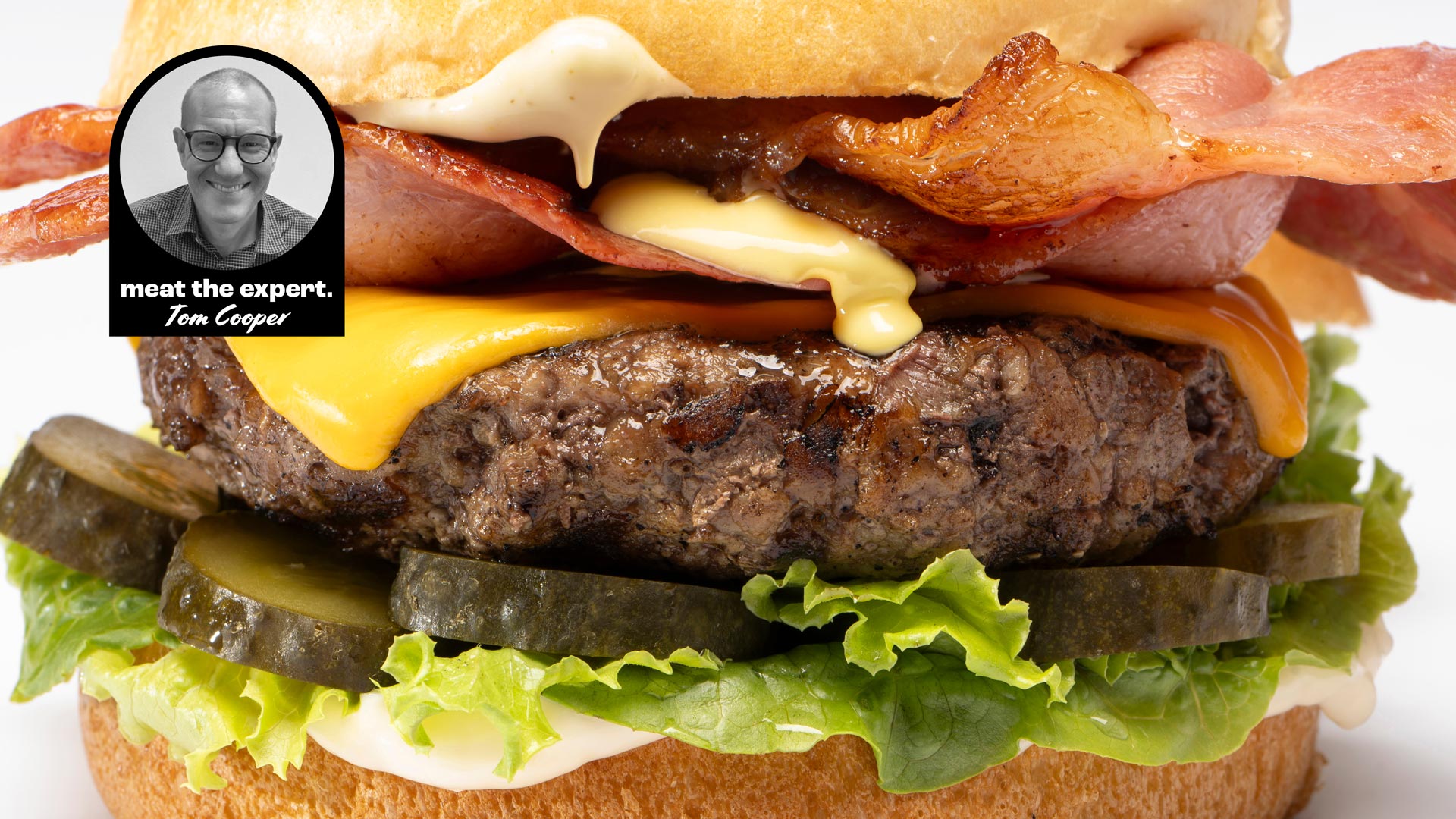 Meat the expert: the perfect hamburger 1 Close-up of a hamburger with lettuce, pickles, a beef patty, melted cheese, bacon, and sauce, with a label featuring Tom Cooper and the text "meat the expert. Tom Cooper."