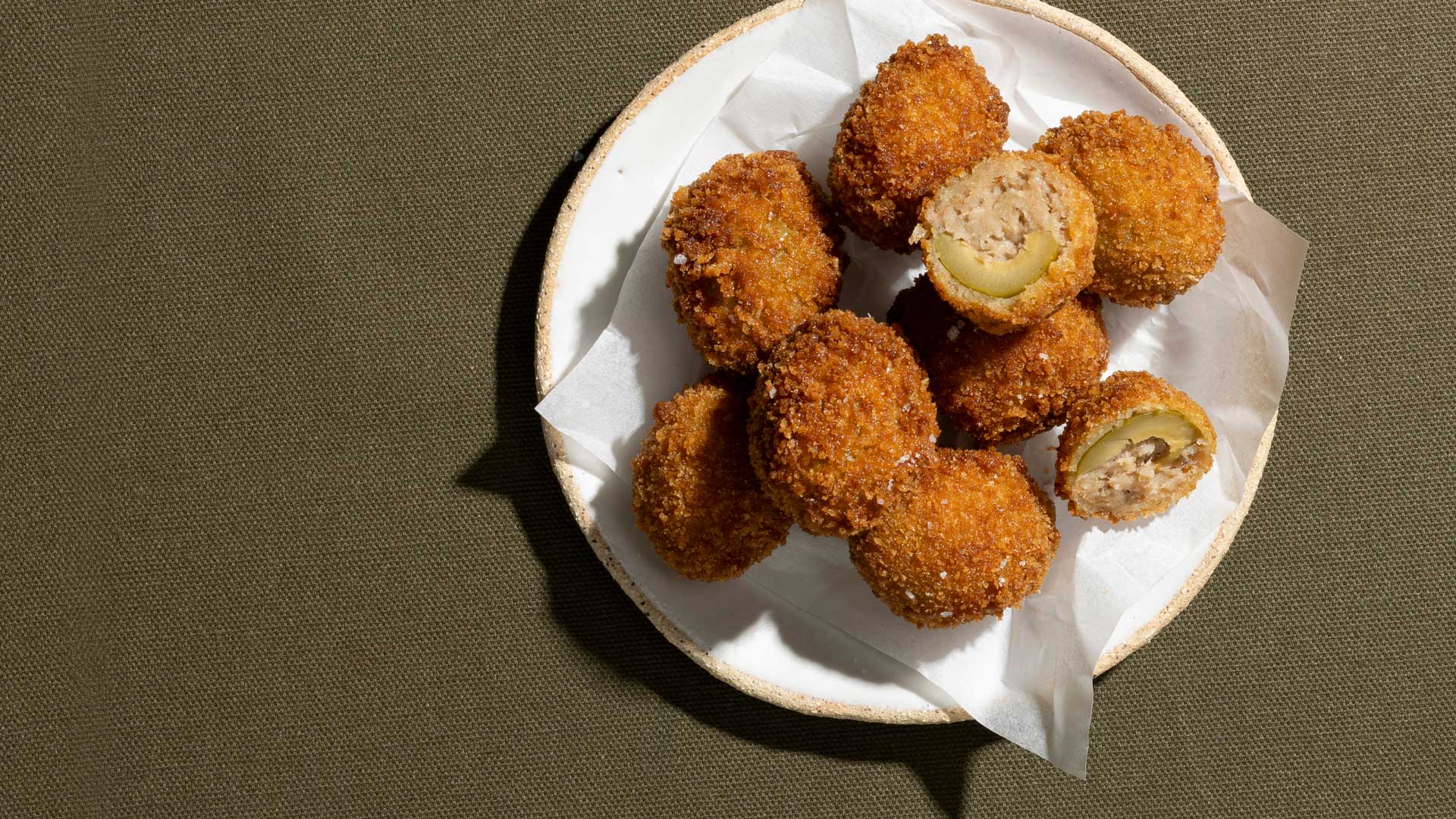 Olive Ascolana 1 A plate of fried stuffed olives, known as Olive all’Ascolana, on a green textured surface. The olives are breaded and fried, with one olive cut in half to show the stuffing inside.